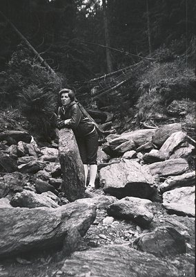 Mid-20th century black-and-white photo of a young woman in rugged forest terrain, gripping a fallen tree trunk for support. S...