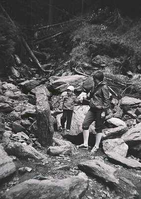 Two hikers navigate rocky terrain in a forested area, likely mid-20th century. The adult assists a child across a stream over...