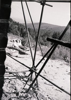Mid-20th century alpine chairlift with rusted metal frame and cables, transporting a seated passenger through forested terrai...