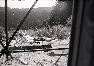 Mid-20th century black-and-white photo of a lone recliner positioned on a gravelly, forested clearing. A person lies relaxed ...