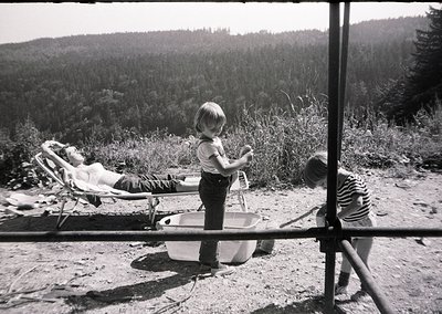 A mid-20th-century black-and-white snapshot captures a lakeside scene: a child in rolled-up pants stands beside a metal washb...