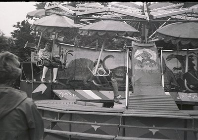 Mid-20th century carnival ride featuring a rotating dinosaur-themed structure with painted murals. Operators in vintage attir...