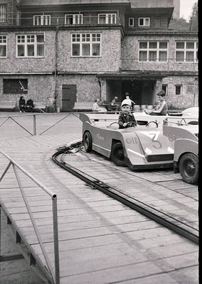 Vintage mid-20th century miniature train ride in a courtyard, featuring a child in a numbered "3" cart labeled "elf" on a tra...