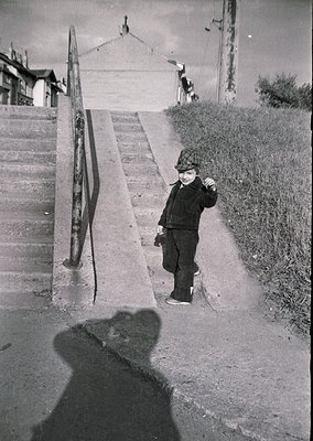 Mid-20th century street scene: Child in winter attire (hat, coat, boots) stands on concrete steps beside a rusted metal raili...