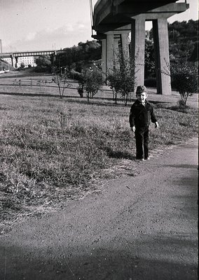 A young boy in mid-20th century attire stands on a paved path beside a concrete overpass, surrounded by overgrown vegetation....