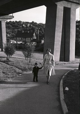 A woman in 1950s-era coat and hat leads a young boy under a modernist concrete overpass, flanked by small trees. Mid-century ...