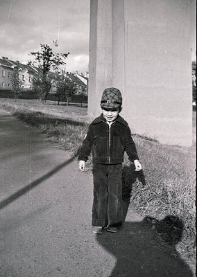 Mid-20th century street scene: Child in 1950s-style jacket and checkered cap holding a small bundle, walking beside a concret...