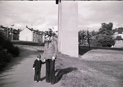 Mid-20th century street portrait: man in 1950s-style jacket and glasses holds child in formal attire beside a tall concrete p...