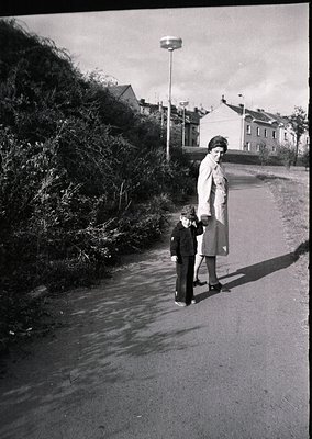 A woman in a long coat and a child in a sweater and shorts walk along a wet, paved road beside dense shrubbery. Mid-20th cent...