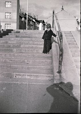 A child in early 20th-century attire descends a wide, concrete staircase flanked by metal railings. Surrounding buildings fea...