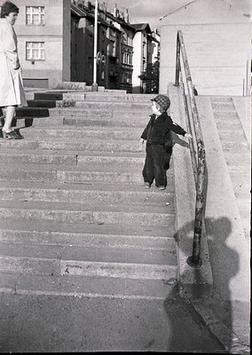Mid-20th century black-and-white street scene: Boy in sweater and cap descends concrete stairs with metal railing, holding ha...