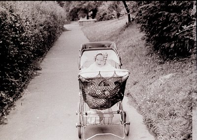 Vintage black-and-white photo of a child in a classic wicker pram on a tree-lined pathway, mid-20th century. Basket details s...