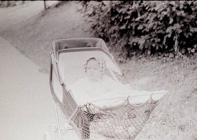 Vintage black-and-white photo of a child reclining in a vintage pram with a mesh canopy, likely mid-20th century. The setting...