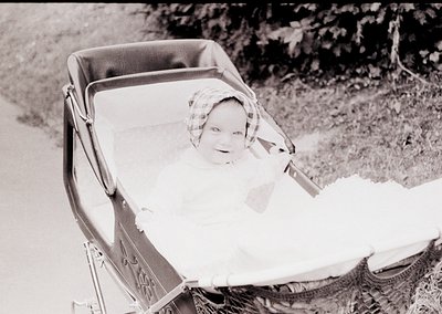Vintage black-and-white photo of a child in a vintage metal-framed stroller, outdoors on a dirt path. The child wears a check...