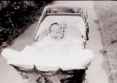 Vintage black-and-white photo of a child wrapped in a thick blanket, seated in a classic wicker pram with a mesh front. The c...