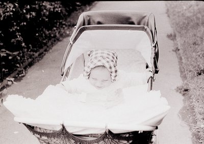 Vintage black-and-white photo of a child in a classic stroller, wrapped in a blanket, with a checkered headscarf. Urban pathw...