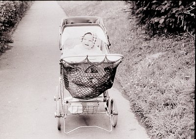 Vintage metal-framed baby carriage with mesh basket, pushed along a paved path beside grassy verge. Infant in a bonnet sits u...