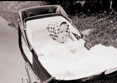Vintage black-and-white photo of an infant in a classic metal-framed pram, wearing a checkered cap. Soft blanket and padded s...