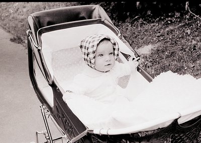 Vintage black-and-white photo of an infant in a classic wooden-framed pram, dressed in a checkered bonnet and long-sleeve gow...