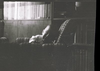 Vintage black-and-white photo of a child resting on a window sill, draped in a striped blanket. Indoor setting with visible b...