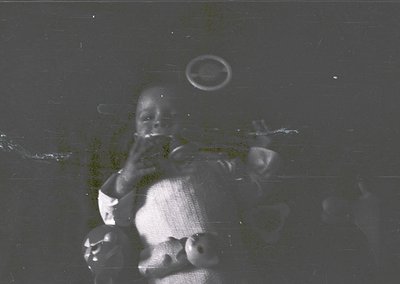 Vintage black-and-white photo of a young child in a seated position, holding a small object to their mouth. The grainy textur...