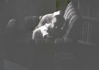 Vintage black-and-white photo of a child sleeping on a plaid blanket, likely mid-20th century. Soft focus and warm lighting s...