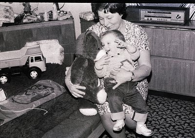 A woman cradles an infant while holding a stuffed animal, likely mid-20th century (1960s–1970s). Surrounding items include a ...