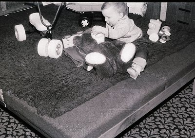 A toddler in mid-1970s medical leg braces plays on a padded hospital bed, surrounded by rolled bandages and a stuffed cow toy...