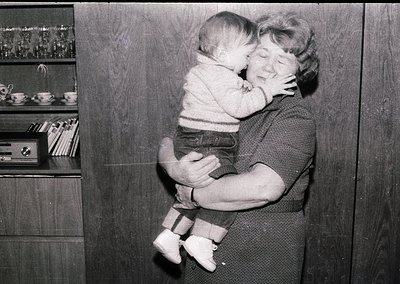 A tender mid-20th-century moment: an adult cradles a baby in a cozy indoor setting. The woman’s sweater and the child’s strip...