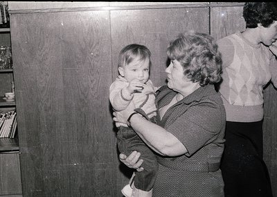 A woman cradles a toddler in a 1960s-70s indoor setting, likely a school or community hall. Wooden cabinets and shelves with ...
