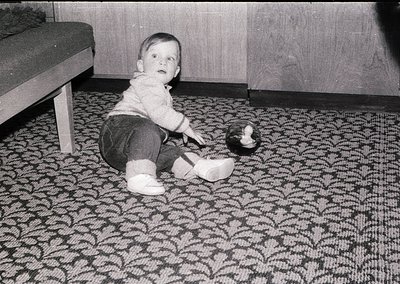 Mid-century indoor scene: toddler in rolled-up trousers and slip-on shoes plays with a small toy duck on a patterned rug, 195...