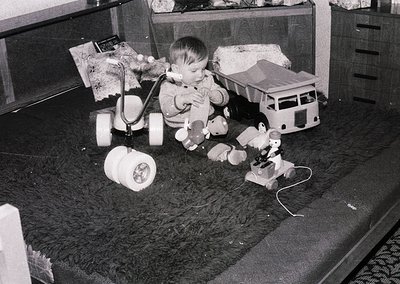 A toddler plays with vintage toys—steam-powered toy train set and toy truck—on a shag-carpeted floor, mid-1960s. Indoor setti...