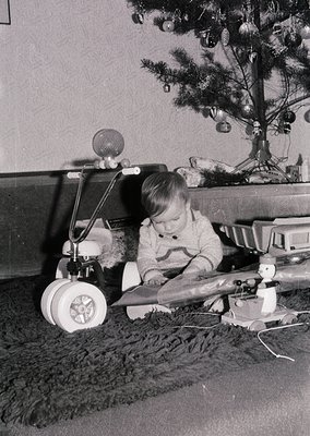 Vintage black-and-white photo of a child playing with a toy airplane on a shag rug, beside a decorated Christmas tree with or...