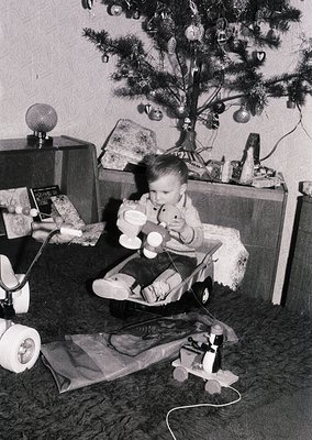 Vintage black-and-white photo of a child in a highchair beside a modestly decorated Christmas tree, holding a toy. Surroundin...