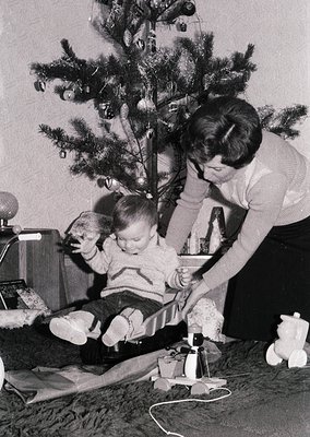 A mid-20th-century (likely 1960s–70s) indoor scene captures a woman decorating a Christmas tree with a child seated on the fl...