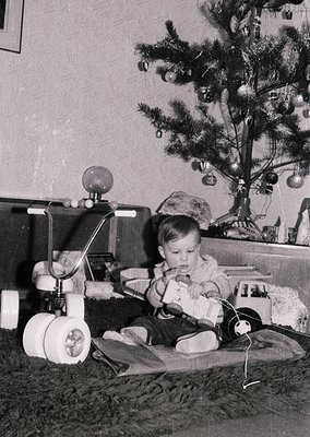 Mid-20th century indoor scene: toddler seated on floor beside vintage toy train set with round wheels and metal track, while ...