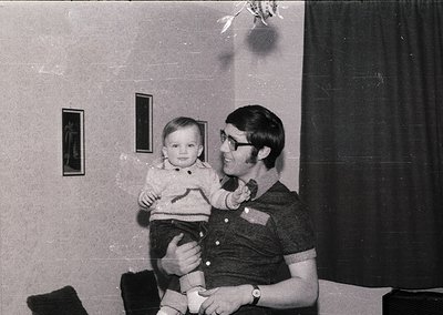 Black-and-white indoor portrait from the 1970s–1980s, featuring a man in a striped polo shirt holding a toddler in a striped ...
