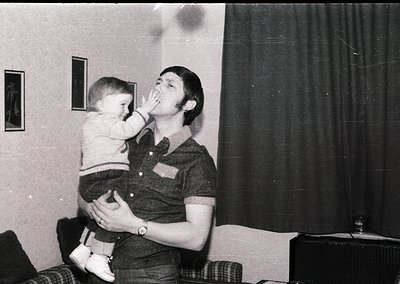 A man in a short-sleeved button-up shirt and jeans playfully kisses a young child indoors, likely mid-1970s. The setting feat...