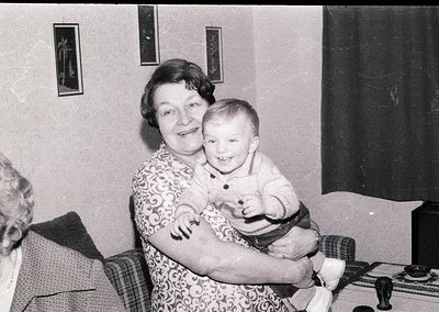 A mid-20th century indoor portrait featuring an elderly woman holding a toddler in a living room setting. The woman wears a f...