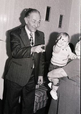Mid-20th century black-and-white photo of an elderly man in a dark suit and patterned tie playfully lifting a toddler in a sw...