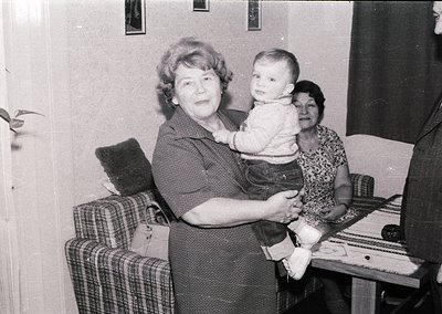 Mid-20th century indoor portrait: woman in 1960s-style dress cradles a baby, flanked by another seated woman in patterned blo...