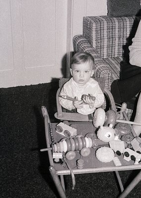 Vintage black-and-white photo of a toddler seated in a highchair, holding a spoon toward a tray filled with classic wooden to...