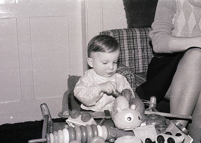 Toddler in 1960s-style sweater plays with vintage toy train set on wooden table. Adult’s plaid skirt and knit sweater visible...