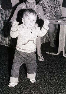 Young child in 1960s-style sweater and knee-high socks, mid-dance pose with arms raised. Indoor setting with patterned sofa a...