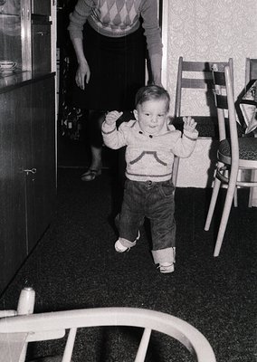 Toddler in mid-stride on dark carpeted floor, wearing 1960s-style sweater and denim pants with white shoes. Adult partially v...