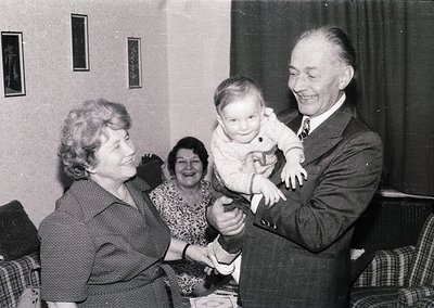 Intergenerational gathering in a mid-20th-century living room, likely 1950s–1960s. Elderly man in suit cradles a baby, while ...
