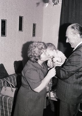 A woman in a dark dress tenderly kisses a child’s forehead while an older man in a suit smiles, holding the child’s hand. Ind...
