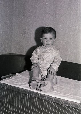 Young child in mid-20th-century attire, seated on a patterned metal-framed cot with a white sheet. Knitted sweater, knee-high...