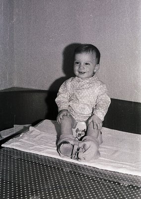 Vintage black-and-white portrait of a toddler sitting on a patterned blanket, wearing a textured sweater and striped socks wi...