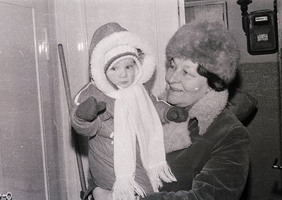 A woman in a fur-lined coat and hat holds a toddler bundled in winter attire, posing indoors near a doorframe. The child wear...